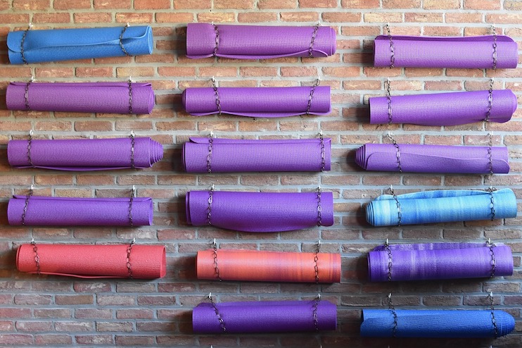Rolled yoga mats in various colors (blue, purple, red, and orange) hanging on a brick wall using metal chains. The mats are neatly arranged in rows and columns, creating a colorful and organized display.