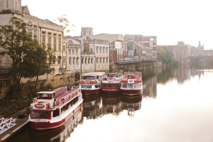 Three red and white passenger boats are moored along the bank of a calm river, adjacent to historic buildings. The sunlight gently illuminates the scene, casting reflections on the water, while trees and architecture line the riverbank in the distance.