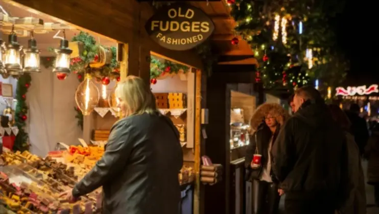 People browsing a festive outdoor market stall that sells fudge, with a sign above reading Old Fashioned Fudges. The stall is decorated with lights and Christmas greenery, and shoppers are enjoying the holiday atmosphere, some holding cups of hot beverages.