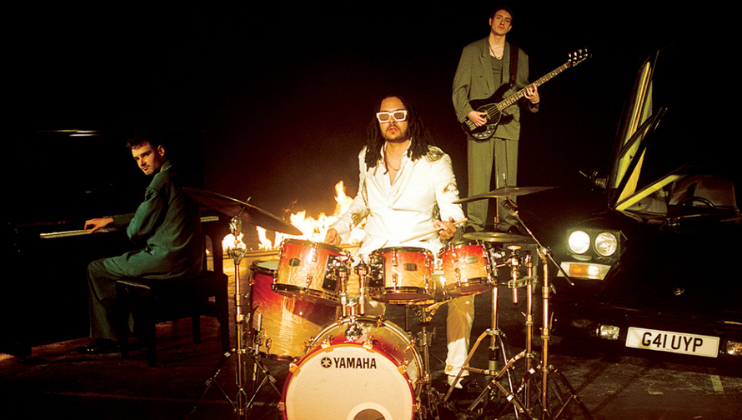 Three musicians are performing: one plays a grand piano, another plays a drum set with flames behind him, and the third stands on a car playing an electric guitar. The car, a black Lamborghini with its doors open, has a license plate reading G41 UYP.